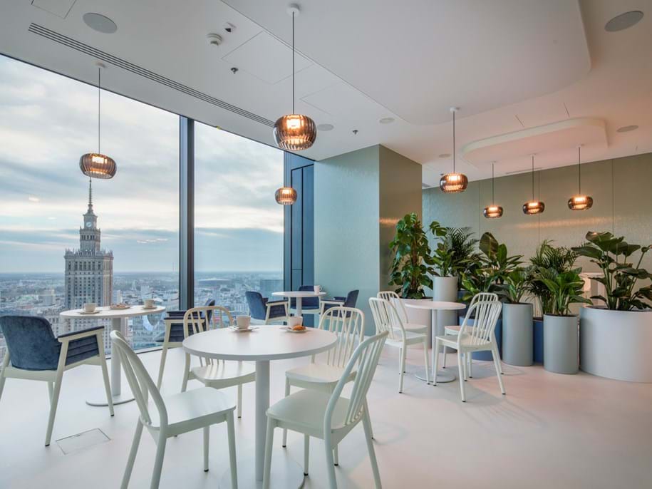 Modernes Bürocafé mit weißen Tischen und Stühlen, umgeben von üppigen Grünpflanzen in großen Töpfen. Bodentiefe Fenster bieten einen Blick auf die Stadtlandschaft mit einem markanten Turm. Von der Decke hängen Pendelleuchten, die das gemütliche Ambiente noch verstärken.