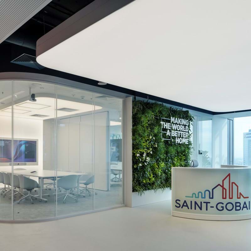Modernes Bürofoyer mit einem Empfangstresen mit dem Saint-Gobain-Logo. Auf der linken Seite befindet sich ein gläserner Besprechungsraum und im Hintergrund ein vertikaler Garten mit dem Slogan Making the World a Better Home. Große Fenster zeigen Gebäude der Stadt.