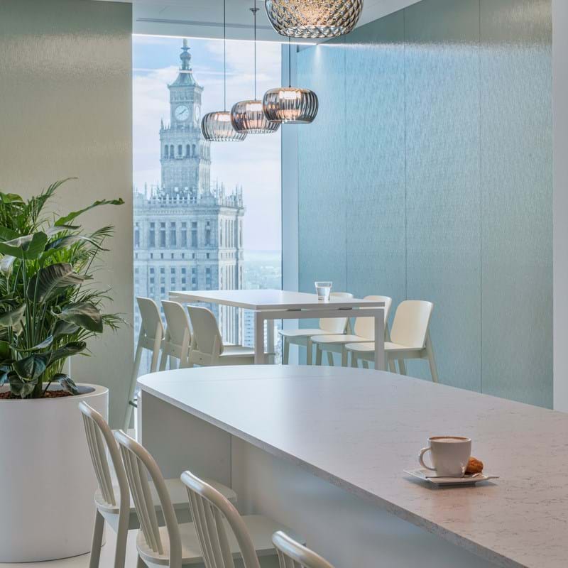Eine moderne Bürolounge mit weißen Stühlen und einem langen Tisch. Hängelampen erhellen den Raum. Große Fenster bieten einen Blick auf einen hohen Uhrenturm und die Stadtlandschaft. Auf dem Tisch stehen eine Tasse und eine Untertasse, und in der Ecke stehen Pflanzen.