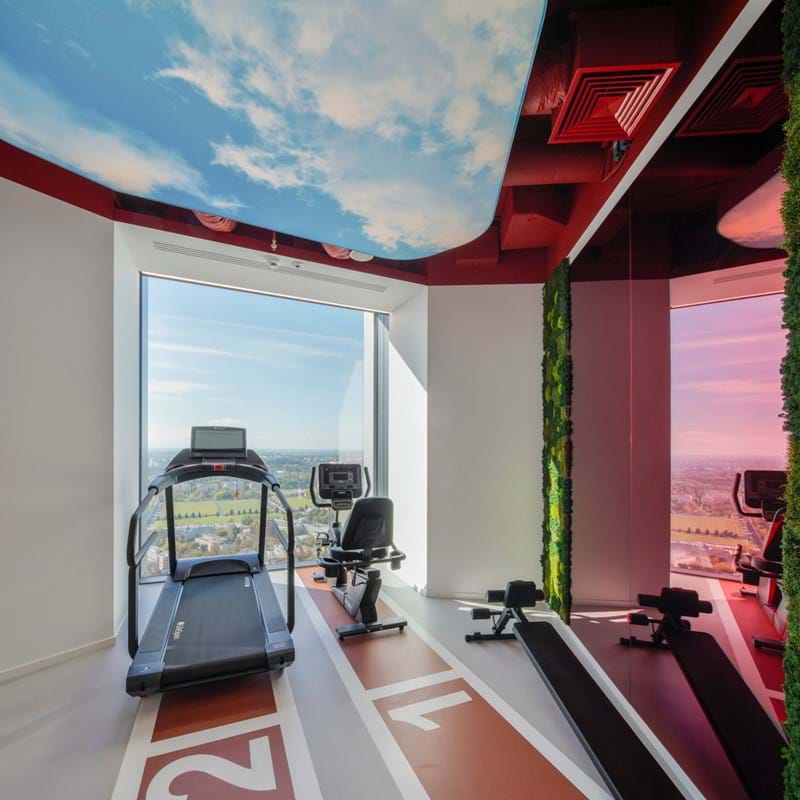 Ein moderner Fitnessraum mit einem Laufband, einem Heimtrainer und einem Rudergerät auf dem Boden einer Leichtathletikanlage. Großes Fenster mit Blick auf eine Stadtlandschaft. An der Decke sind Himmel und Wolken abgebildet, und an den Wänden gibt es grüne Akzente.