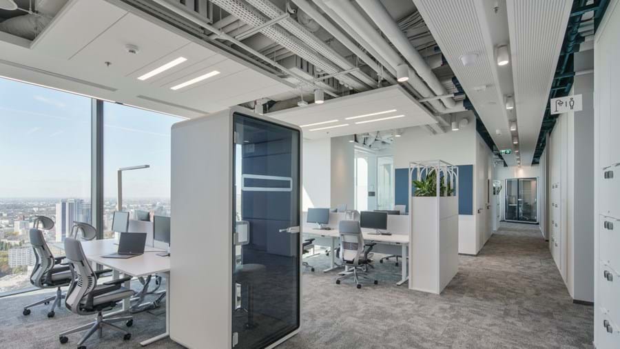 Eine moderne Büroeinrichtung mit mehreren Arbeitsplätzen mit Computern und Stühlen. Der Raum ist offen mit einem raumhohen Fenster, das eine Stadtlandschaft zeigt. An der Decke sind freiliegende Rohrleitungen zu sehen, und es gibt Topfpflanzen und eine Telefonzelle.