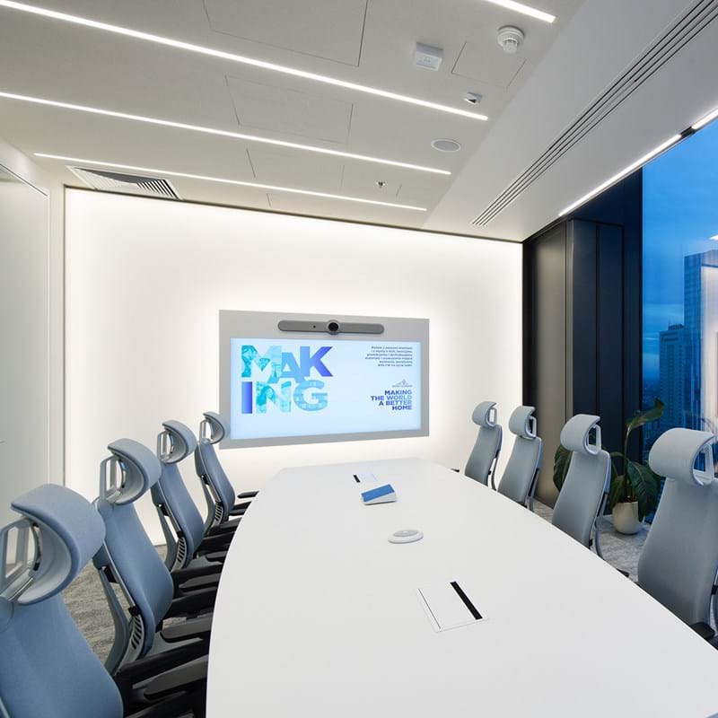 Ein moderner Konferenzraum mit einem langen weißen Tisch und zehn ergonomischen Stühlen. Ein großer Bildschirm an der Wand zeigt farbige Texte an. Vom Boden bis zur Decke reichende Fenster bieten einen Blick auf eine Stadtlandschaft. Der Raum ist hell erleuchtet, die Wände sind weiß und der Boden mit Teppich ausgelegt.