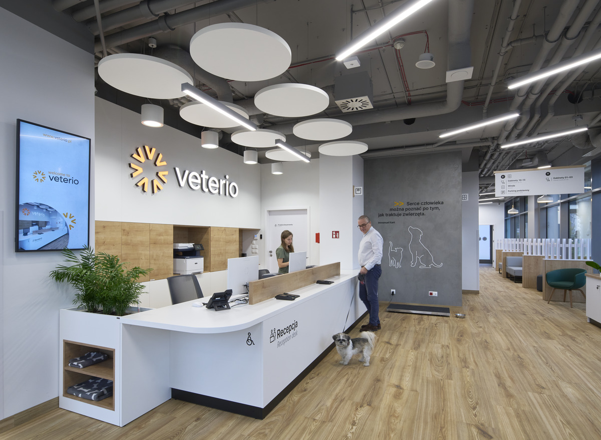 Eine moderne Tierklinik mit einem Empfangstresen. Eine Frau steht hinter dem Tresen, während ein Mann mit einem angeleinten Hund davor steht. Der Raum ist mit Holzfußböden, Deckenleuchten und Wandmalereien mit Tiermotiven ausgestattet.
