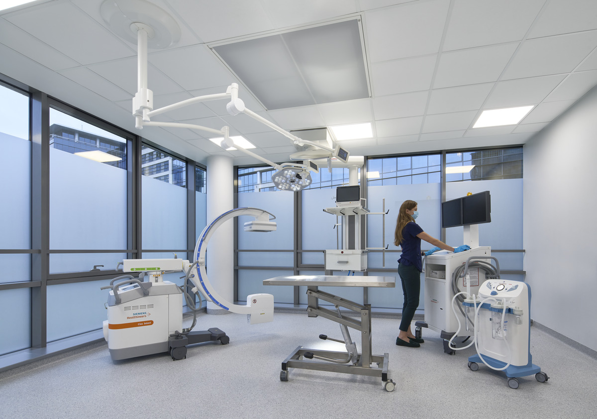 Ein moderner medizinischer Raum mit großen Fenstern, in dem sich moderne Geräte wie ein Bildgebungsgerät, ein Operationstisch und Monitore befinden. Eine Person in einem marineblauen Hemd bedient eine Maschine, über der eine helle Beleuchtung hängt.