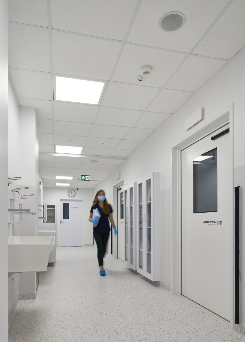 Eine Person, die eine Maske trägt, geht einen sauberen, gut beleuchteten Krankenhausflur entlang. Der Flur ist mit Waschbecken und Schränken zum Händewaschen ausgestattet. An den Türen befinden sich Schilder und an der Wand eine Uhr, die die Uhrzeit anzeigt.