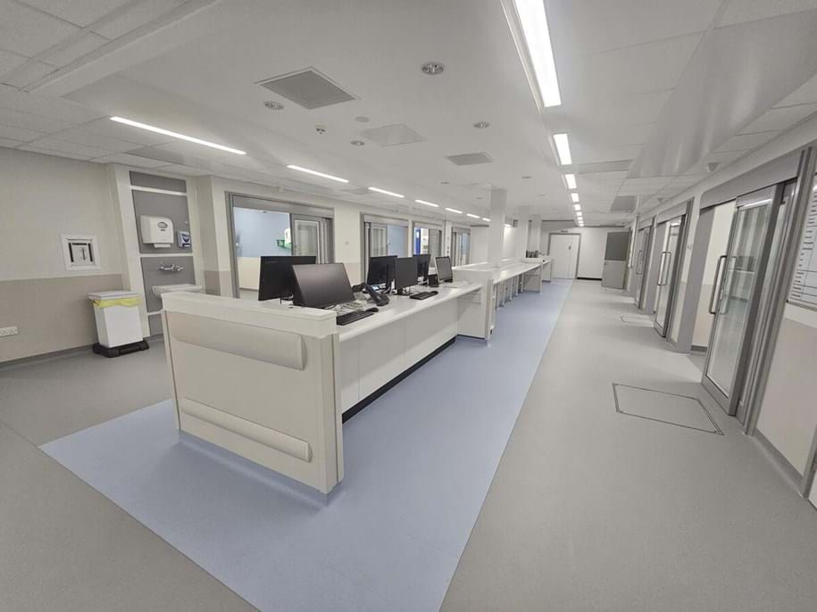 A modern, empty hospital ward with a central nurses' station featuring several monitors, surrounded by patient rooms with glass doors. The area is brightly lit and clean, with light-coloured floors and walls.