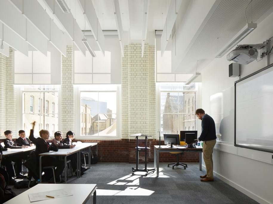 A modern classroom with several students seated at desks facing a teacher, who stands near a whiteboard. The room is well-lit with large windows, white walls, and exposed brick. The students appear attentive and engaged.