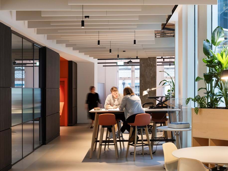 Modern office space with three people; two are seated at a high table working together, while one person walks by in the background. Large windows and indoor plants create a bright, open atmosphere.