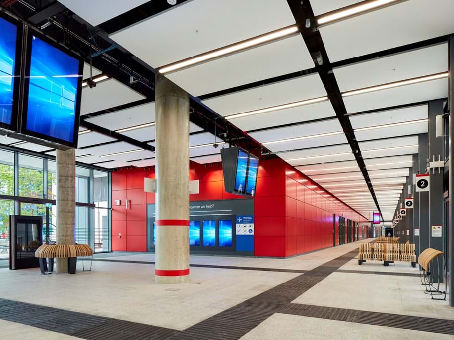 Modernes Busbahnhof-Interieur mit hohen Decken, roten und grauen Wänden, digitalen Informationsbildschirmen und Holzbänken. Helle Beleuchtung erhellt den Raum, im Hintergrund ist ein langer Korridor zu sehen. Schilder mit Nummern hängen von der Decke. Auf der linken Seite sind große Eingangstüren zu sehen, durch die Tageslicht in den Terminal fällt.