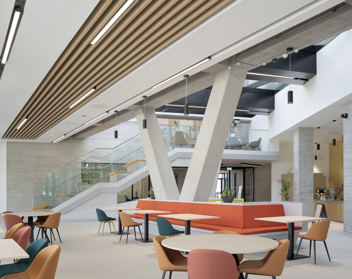 Modern interior space with high ceilings and natural light, featuring tables and colourful chairs. Acoustic ceiling baffles complement the V-shaped concrete pillars, and a staircase with glass railings to enhance the contemporary design.