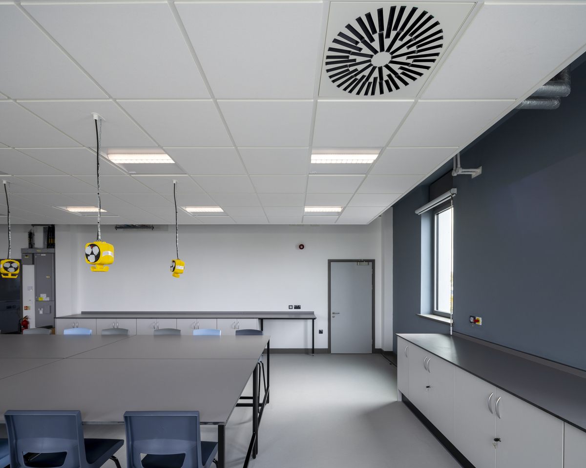 A modern science classroom with grey tables, blue chairs, white ceiling tiles, large ventilation fans, yellow electrical sockets hanging from the ceiling, and cupboards along the walls. A window and door are visible to the right.