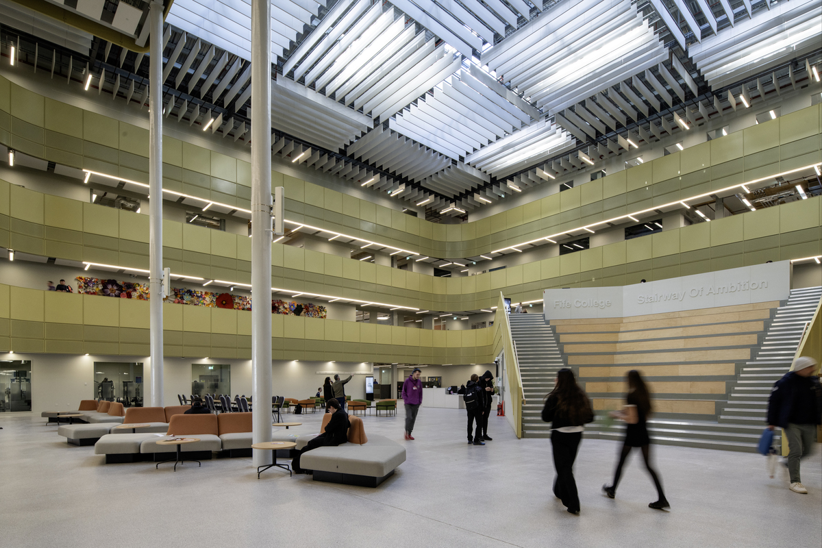 Spacious modern atrium with skylights, tiered seating, and people walking or sitting on benches. Multiple floors are visible, with colourful art on a wall and open spaces throughout.
