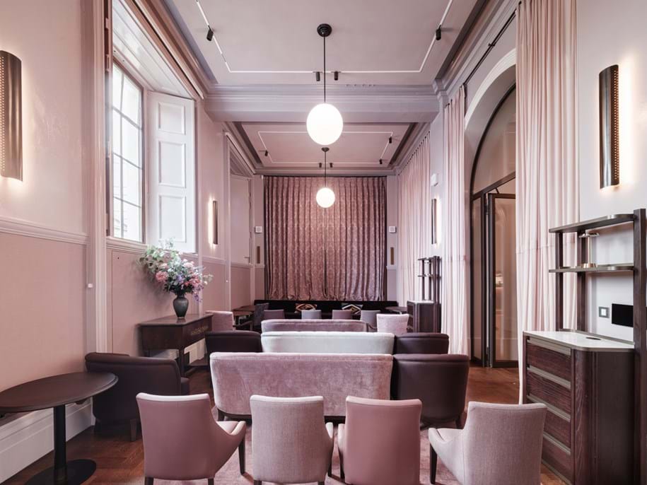 A stylish, elegant lounge with pastel pink and mauve seating, wooden floors, tall windows, soft lighting, and modern décor. A floral arrangement sits on a side table, and curtains frame an arched doorway at the back.