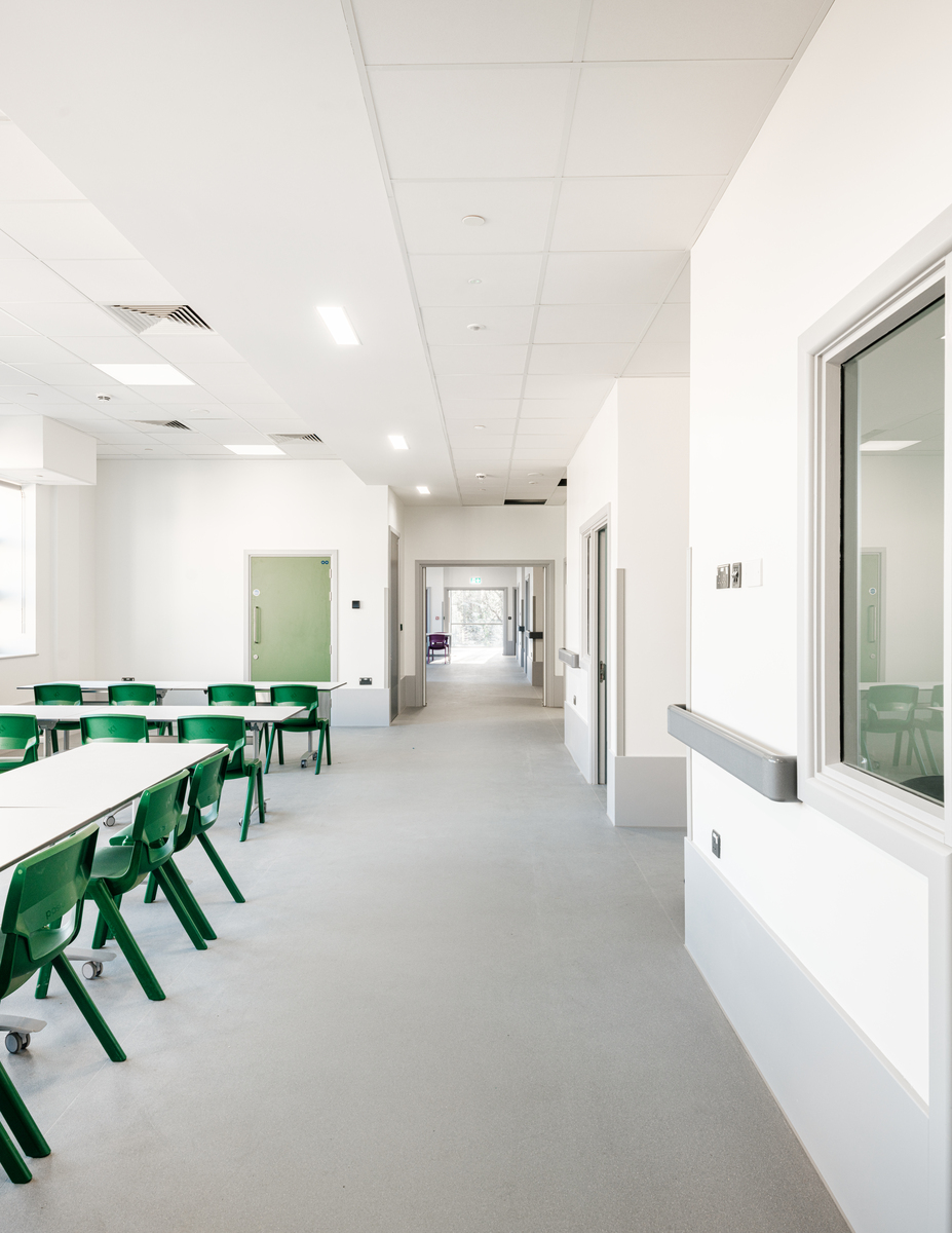 A bright, modern corridor in a school or institutional building, with green chairs around tables on the left and wide windows on the right; the corridor extends towards distant doors.