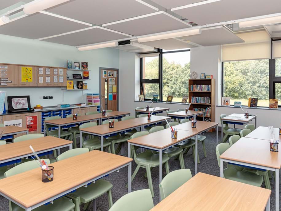 A bright, tidy classroom with several desks and green chairs, each desk holding a pencil pot. There are bookcases, wall displays, windows with views of trees, and educational materials around the room.