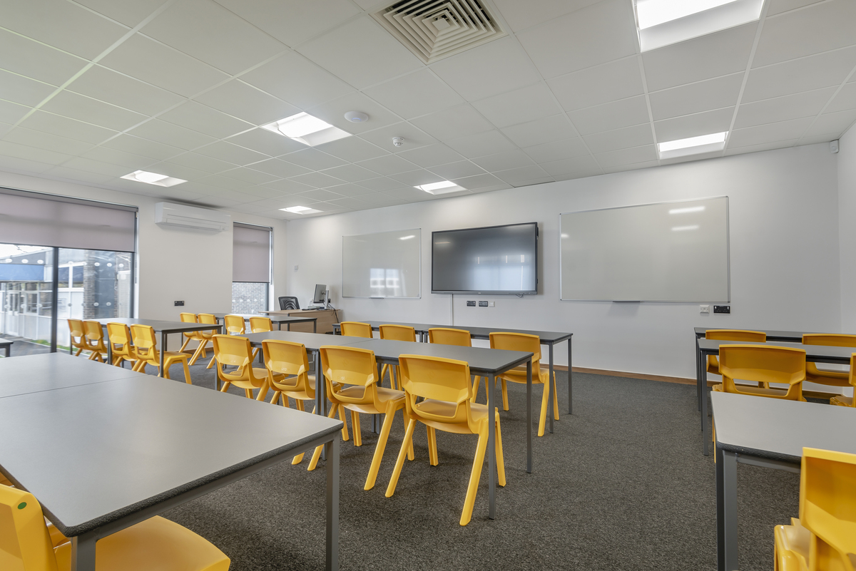 A modern classroom with a whiteboard and digital screen on the wall, multiple rows of yellow chairs and desks facing forward, and large windows allowing natural light to fill the room. The white acoustic ceiling features integrated lighting. The setting appears to be ready for an educational session or lecture.
