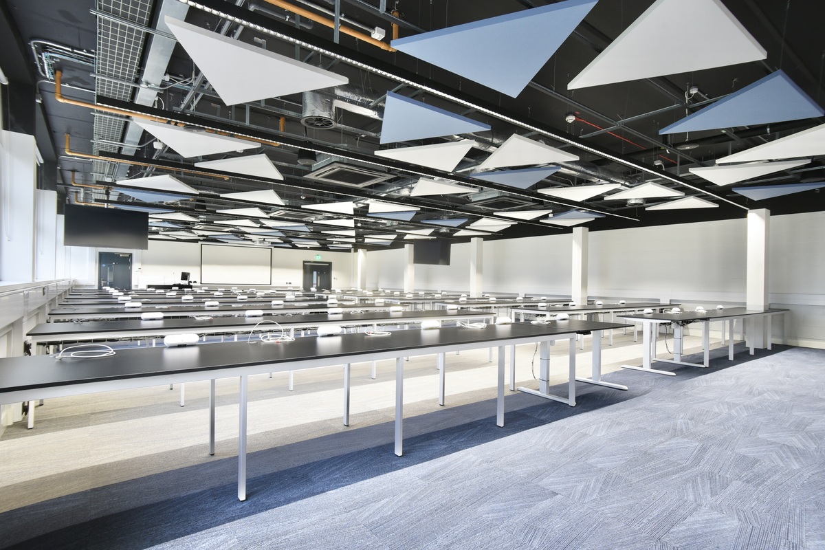 A modern, spacious classroom with long black desks, white chairs, and triangular acoustic panels on the ceiling. The room is brightly lit with large windows and has a clean, minimalist design.