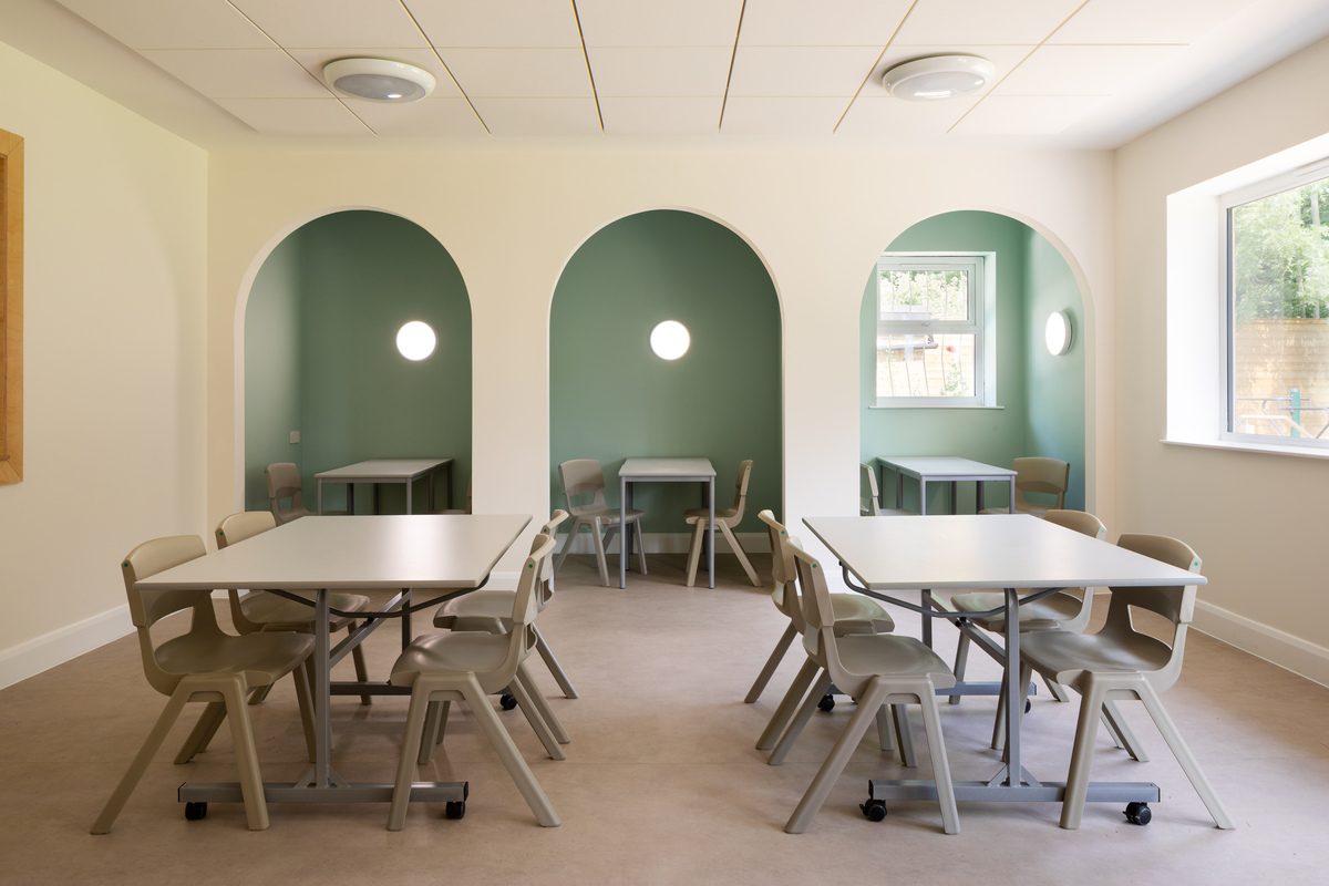 A bright, modern classroom with beige walls and green alcoves, each containing a table and chairs. Four square tables with beige chairs are arranged in the centre, and sunlight streams in from a large window on the right.