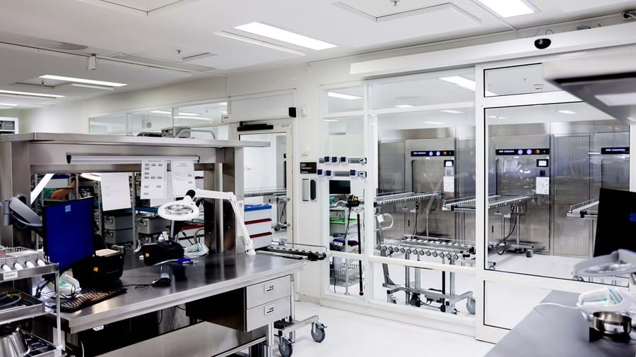 Et moderne, rent laboratorium med arbejdsstationer i rustfrit stål, computere og videnskabeligt udstyr. Store glasvinduer adskiller laboratoriet fra et andet rum med industrimaskiner og transportbånd. Klar loftsbelysning.