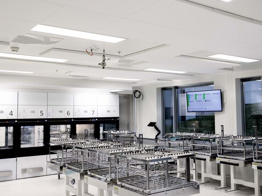 A modern, bright laboratory with multiple empty metal racks, large windows, a wall-mounted monitor displaying data, and numbered equipment units along the back wall. The room appears clean and organised.