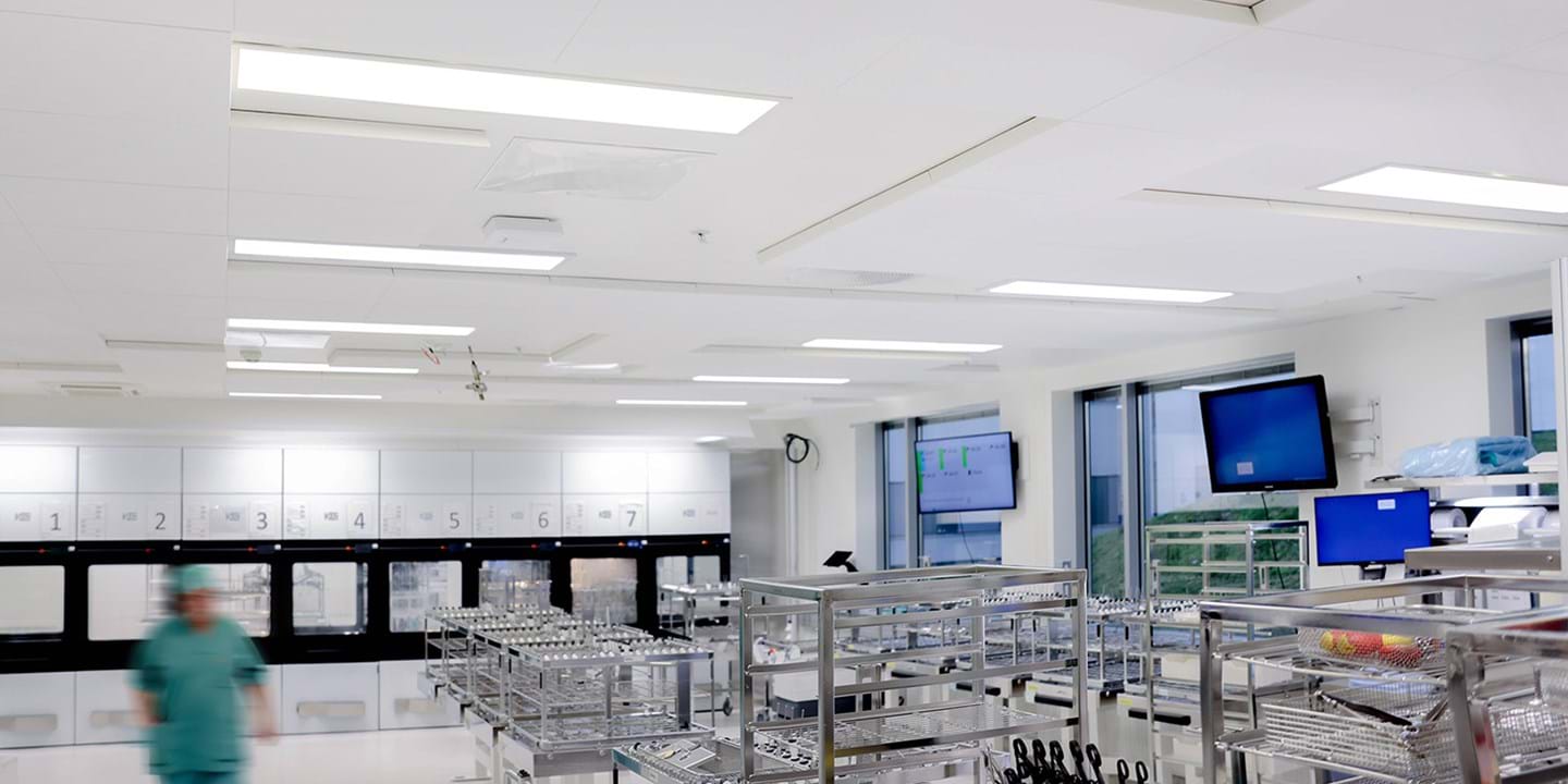 A modern, sterile hospital sterilisation room with metal racks holding surgical instruments. A person in green scrubs walks through the bright, clean space. Monitors and cupboards line the white walls.