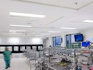 A modern, sterile hospital sterilisation room with metal racks holding surgical instruments. A person in green scrubs walks through the bright, clean space. Monitors and cupboards line the white walls.