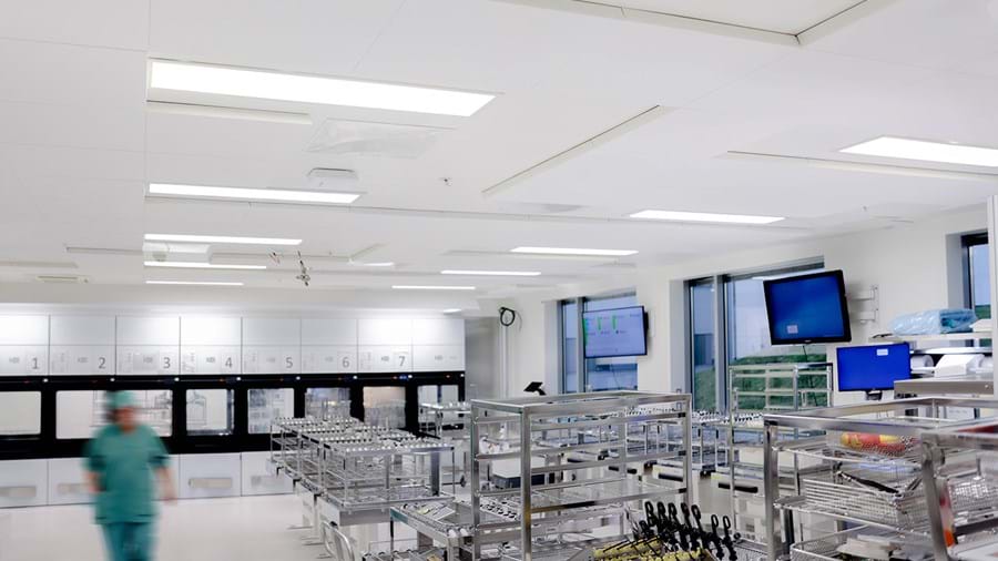 Et moderne, sterilt hospitalsteriliseringsrum med metalstativer til kirurgiske instrumenter. En person iført grøn operationsuniform går gennem det lyse og rene rum. Skærme og skabe er placeret langs de hvide vægge.