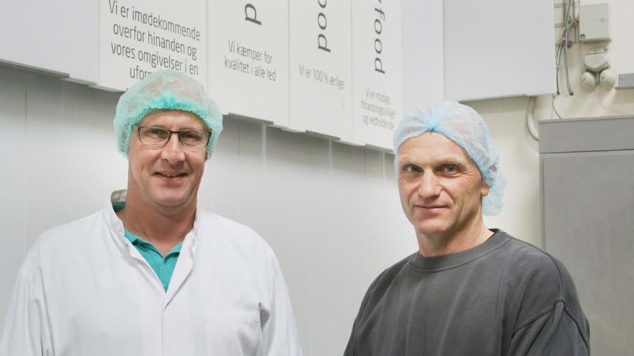 Zwei Männer mit Haarnetzen und Arbeitskleidung stehen in einem Innenraum vor weißen Schildern mit Text. Einer trägt eine Brille und einen Laborkittel, der andere ein dunkles Hemd.