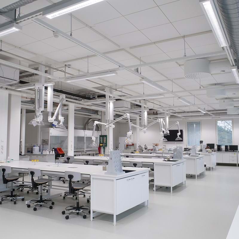 Et lyst og moderne laboratorium med hvide borde, taburetter, loftsbelysning og forskelligt videnskabeligt udstyr. Rummet er rent, rummeligt og for det meste tomt, med store vinduer, der lukker naturligt lys ind.