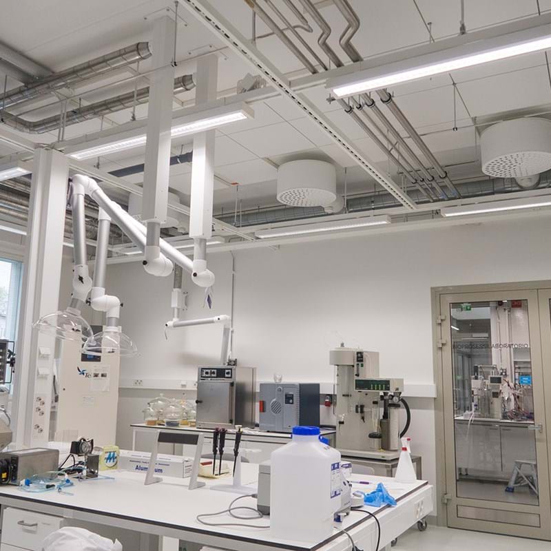 Et moderne laboratorium med hvide vægge og arbejdsbænke, laboratorieudstyr, ventilationskanaler i loftet og videnskabelige apparater. Store vinduer lukker naturligt lys ind, og en glasdør fører til et andet laboratorium.