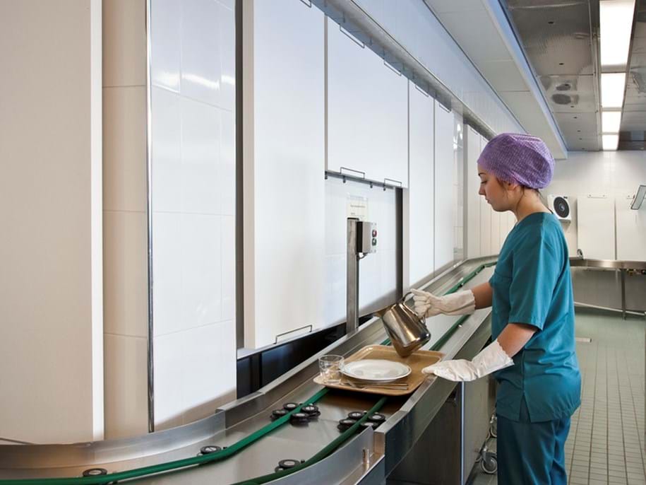 A person in green scrubs and a hair net places a meal tray onto a conveyor belt in a clean, modern hospital or cafeteria kitchen.