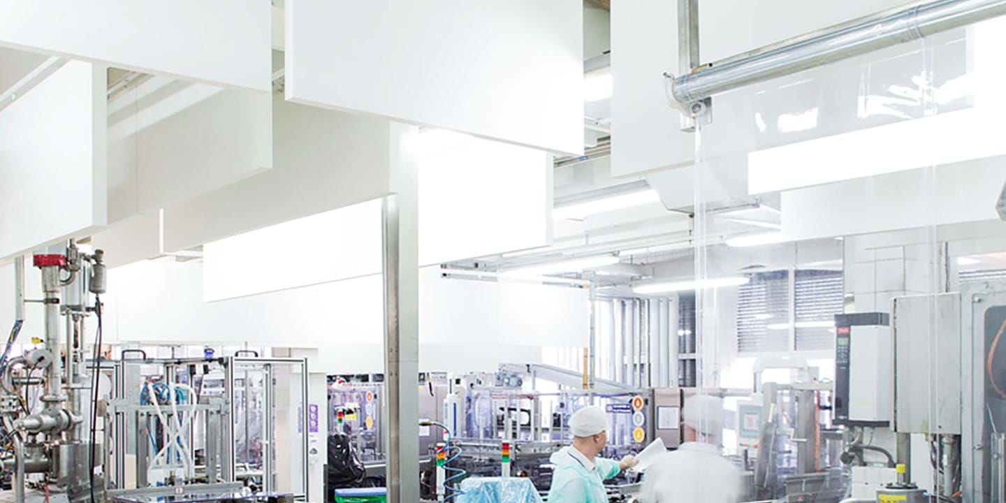 A bright, clean industrial facility with workers in protective clothing operating machinery and equipment, surrounded by stainless steel surfaces and large overhead panels.