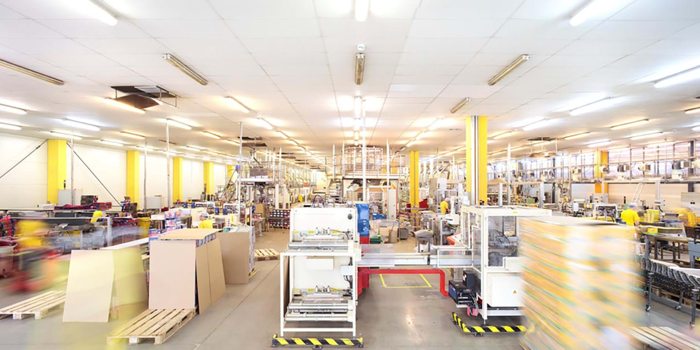 Bright, spacious factory floor with high ceilings, fluorescent lights, yellow pillars, machinery, cardboard boxes, pallets, and blurred figures in motion, indicating an active production or packaging environment.
