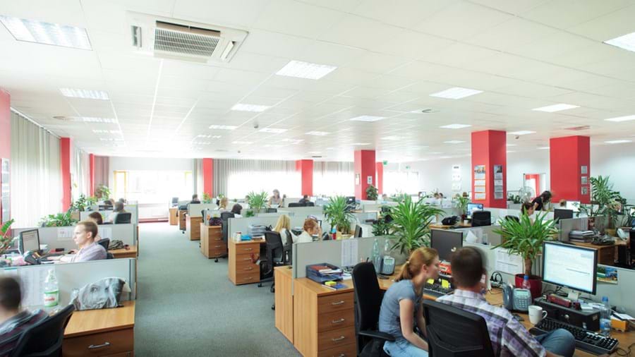 A modern, spacious office with rows of desks and computers, several people working, large windows letting in natural light, red pillars, and green plants throughout the workspace.