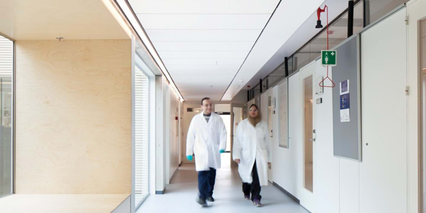 Kaksi valkoisiin laboratoriotakkeihin pukeutunutta henkilöä kävelee laboratorion tai lääketieteellisen laitoksen valoisaa, modernia käytävää pitkin. Käytävän toisella puolella on suuret ikkunat ja toisella puolella ovet, joissa on kylttejä.