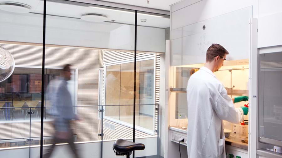Valkoiseen laboratoriotakkiin pukeutunut tutkija työskentelee modernissa laboratoriossa vetokaapin ääressä, samalla kun toinen henkilö kävelee ohi lasiseinän ulkopuolella. Tutkijan lähellä laboratorion lattialla on musta jakkara.