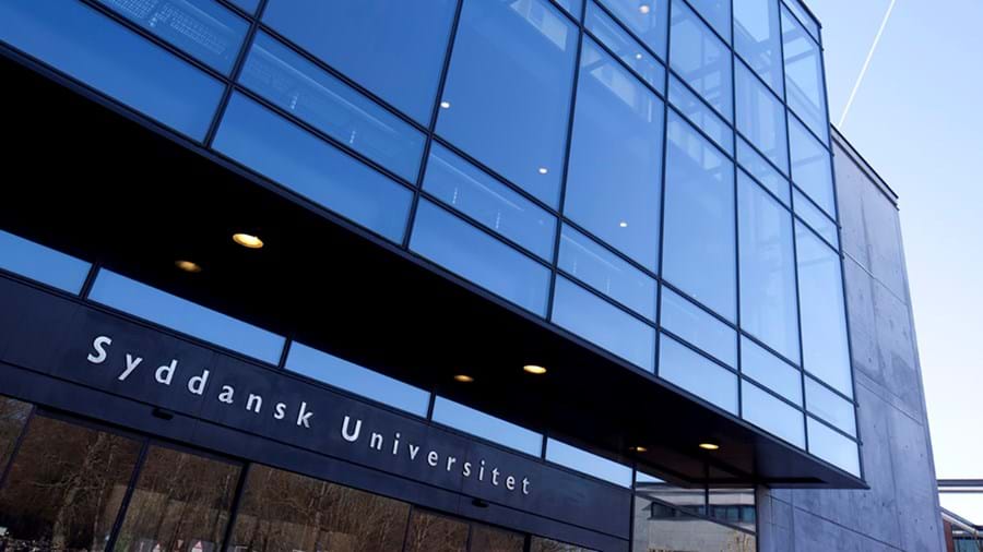 Moderne Glasfassade eines Gebäudes mit dem Schild „Syddansk Universitet“ über dem Eingang, in dem sich der Himmel und die Umgebung spiegeln.
