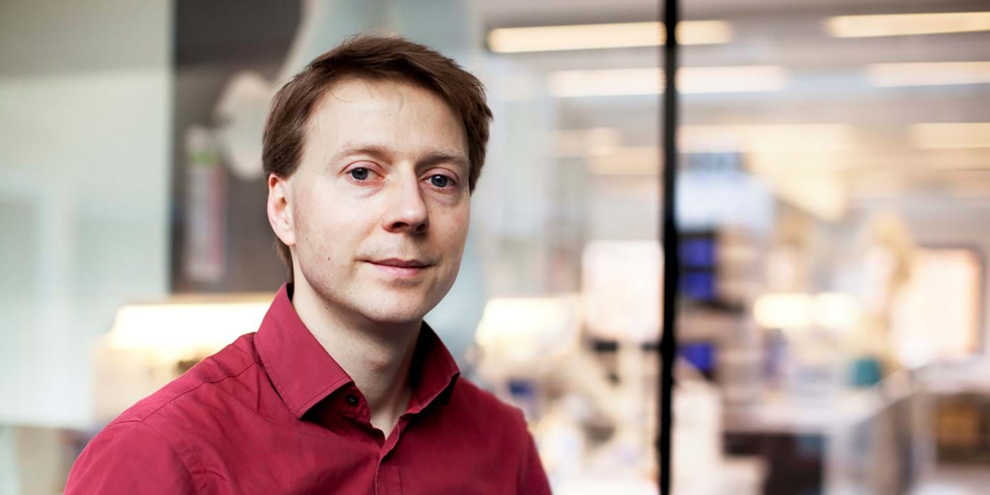 Ein Mann mit hellbraunem Haar und einem roten Hemd steht in einem Innenbereich vor einer Glaswand. Im Hintergrund ist eine verschwommene moderne Büro- oder Laborumgebung zu sehen.