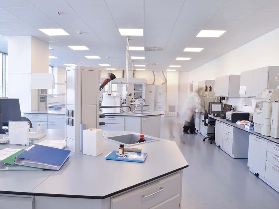 A bright, modern laboratory with white walls and workstations, scientific equipment, computers, and shelves. A person in a lab coat is blurred, walking in the background. Large windows let in natural light.