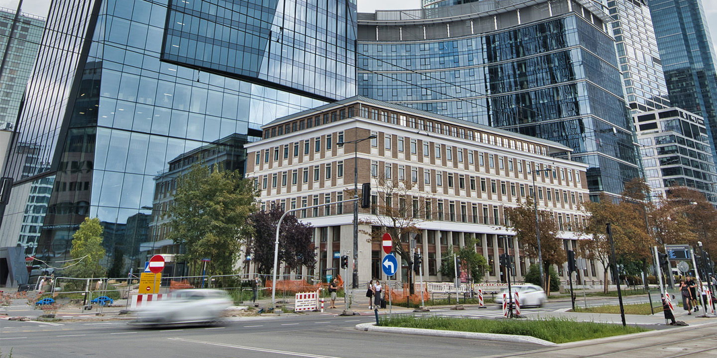 A historic rectangular building with columns stands among modern glass skyscrapers; trees, cars, and street signs are visible in the foreground.
