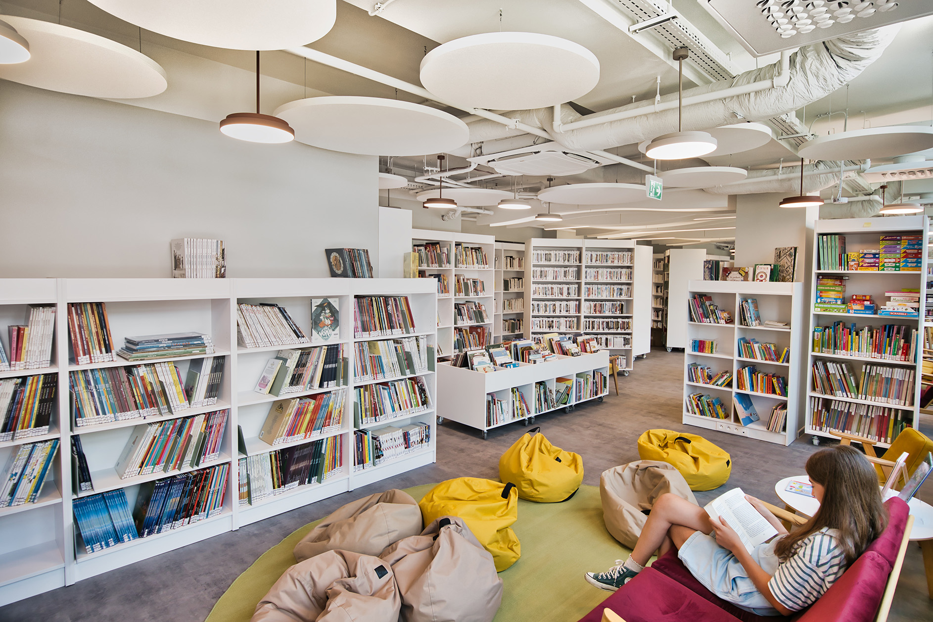 A modern library with white bookshelves, colourful books, round ceiling lights, bean bags on a green rug, and a girl sitting and reading a book.