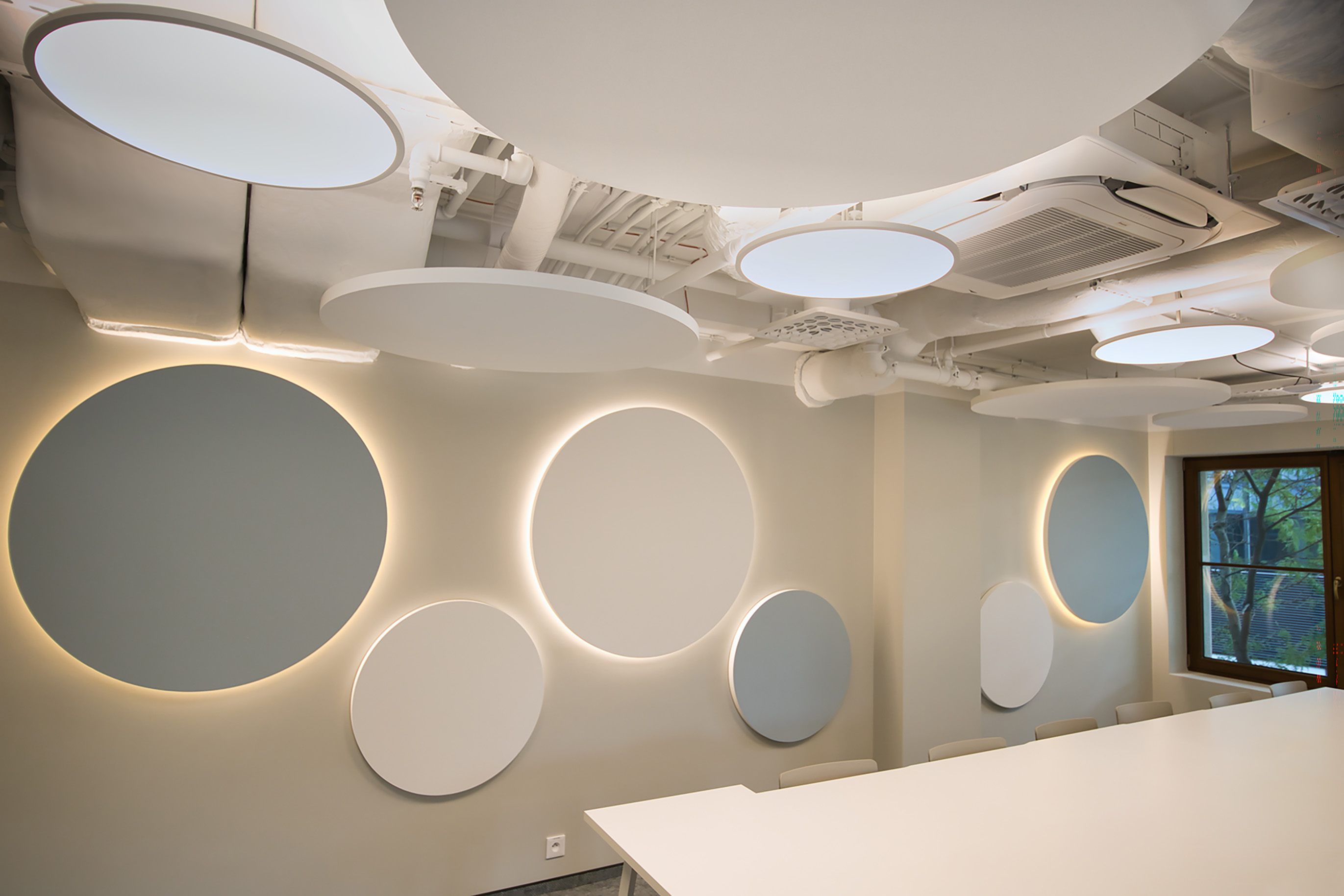 Modern interior with large circular panels on the wall, some illuminated, and round ceiling lights. The space has neutral colours, exposed ducts, and a large window showing greenery outside. Minimalist, clean, and bright design.