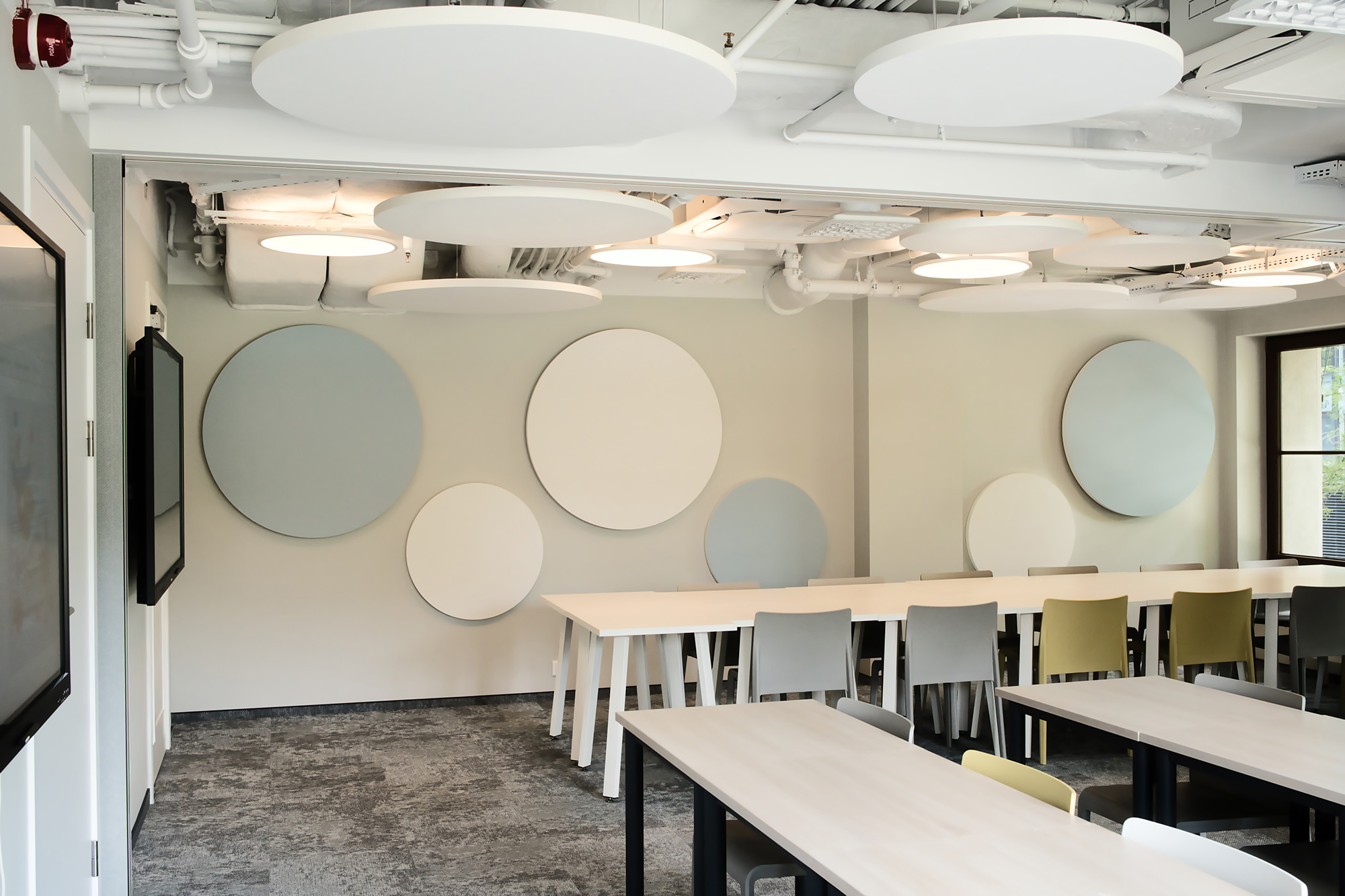 Modern classroom with white tables, mixed white and green chairs, and large circular acoustic panels on the walls and ceiling. The room is bright with natural light from windows.