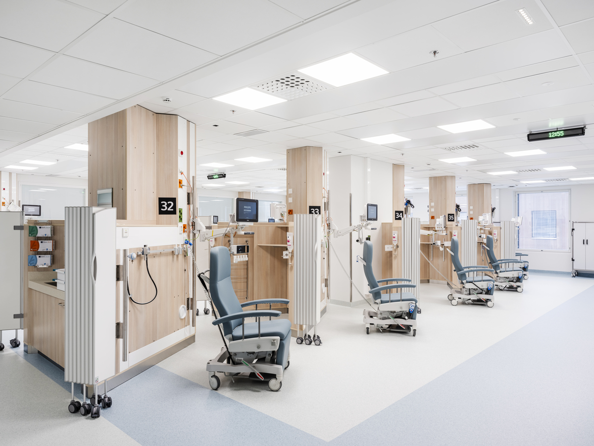 Bright, modern hospital treatment area with several reclining medical chairs separated by wooden partitions, medical equipment, and monitors. The space is clean, organised, and well-lit, with white walls and large overhead lights.