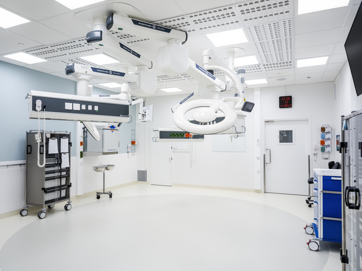 A modern, empty hospital operating theatre with bright lighting, advanced surgical equipment hanging from the ceiling, medical trolleys, and a clock on the wall displaying the time 07:36. The room is clean and organised.
