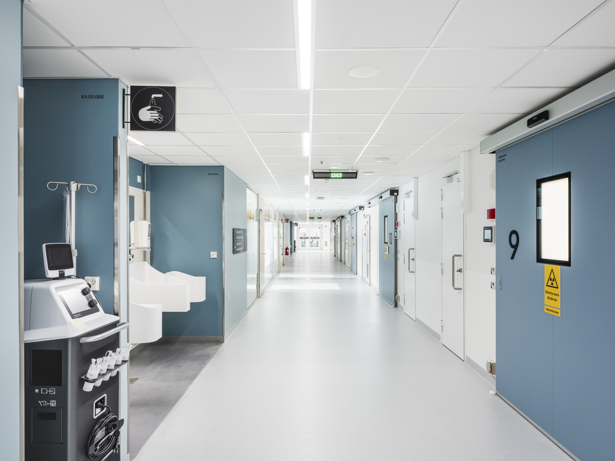 A clean, modern hospital corridor with bright lighting, blue and white walls, numbered patient room doors, medical equipment, and handwashing basins along the sides. The corridor appears empty and well-maintained.