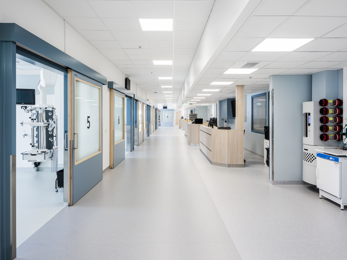 A bright, modern hospital corridor with numbered patient rooms on the left, a central nurses' station, white and blue walls, and medical equipment neatly arranged along the corridor.