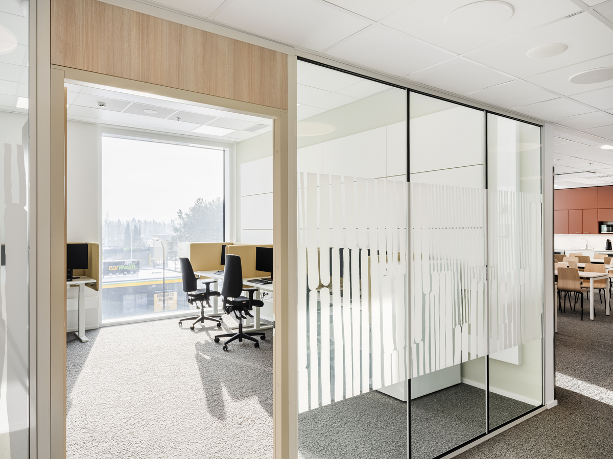 Bright modern office space with glass walls, frosted decorative patterns, workstations with computers and chairs near a large window, and a communal area with tables and chairs in the background.