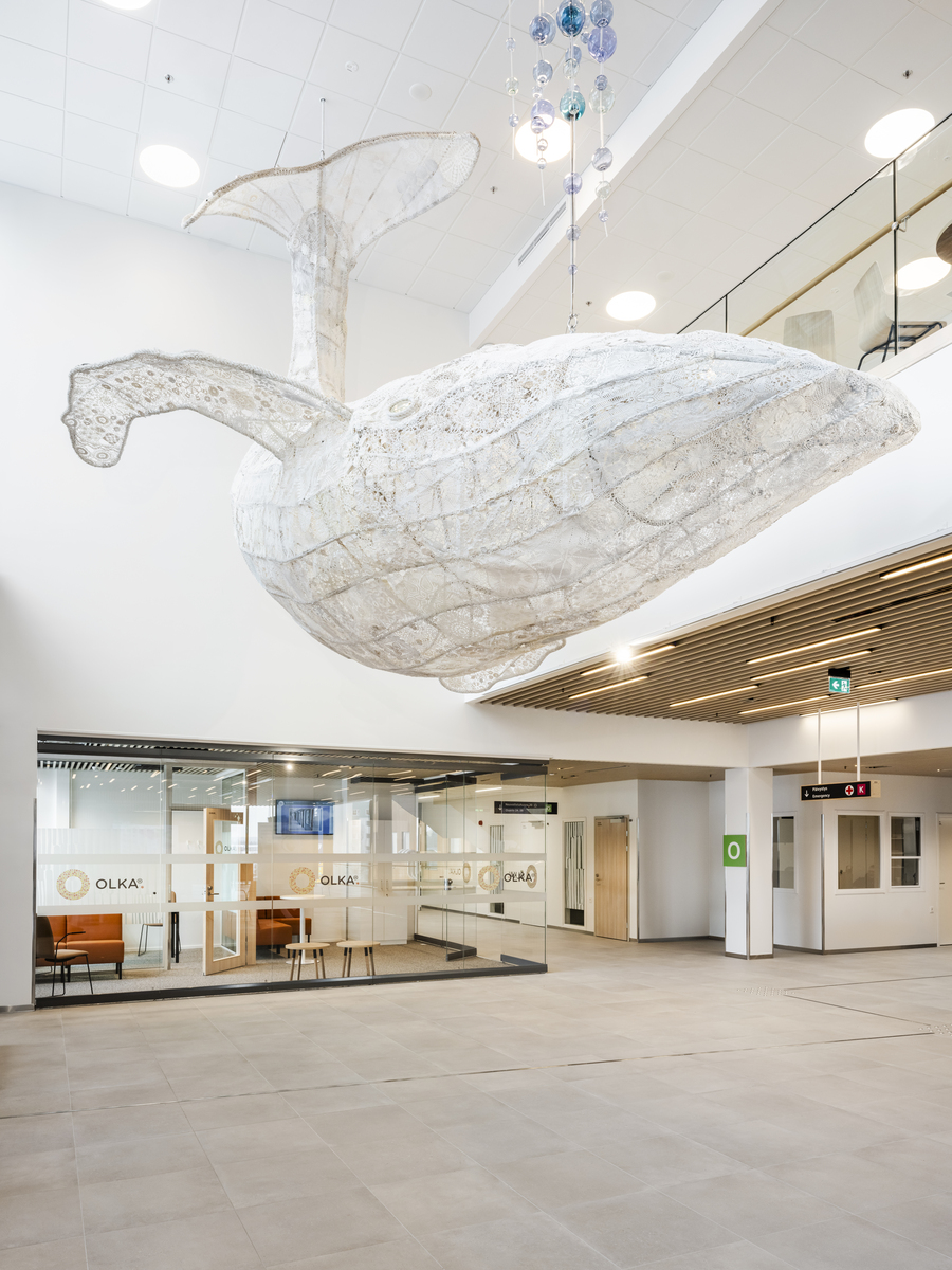 A large, white, whale sculpture hangs from the ceiling in a modern, spacious building lobby with glass-walled offices and minimalist décor.