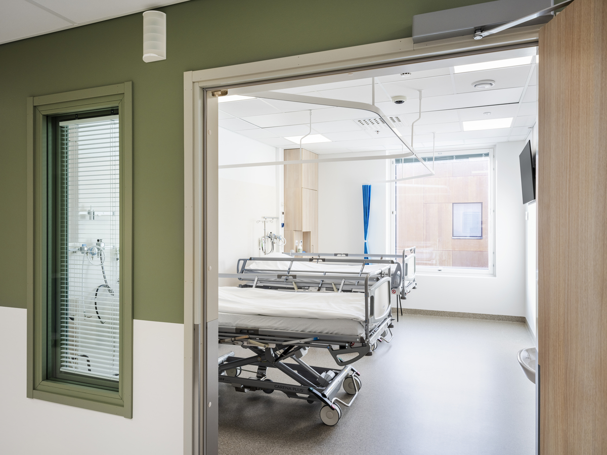 A modern, empty hospital ward with a single bed, medical equipment, and a large window letting in natural light. The door is open, and a green-framed window is visible in the corridor outside.
